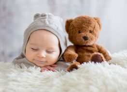 baby boy dressed in overall with a toy bear on bed