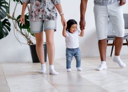 Positive little one and half year mixed-time girl walks around house and takes her first steps holding hands of father and mother in cozy home