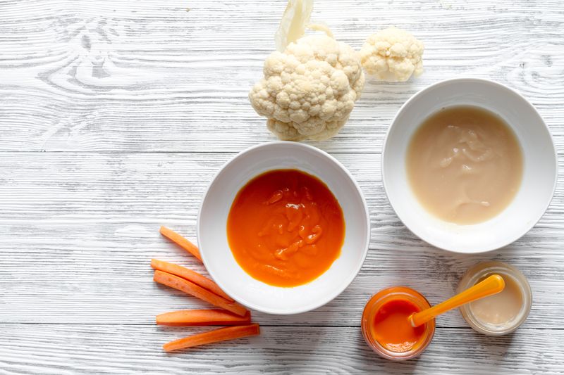 Two bowls of vegetable puree, two jars of puree and a silicone baby spoon