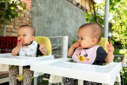 twin babies, one with a colorful polka dot fruit bib and another with a pink bear bib, eating fruits on white high chairs