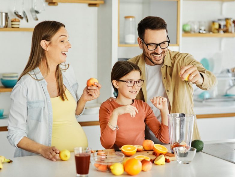 what to buy for second baby - a happy and pregnant mom, dad, and their daughter making orange juice in the kitchen