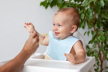 when do babies need bibs - shot of a caucasian baby wearing a blue bib, sitting on his high chair, and smiling at his parent while they feed him