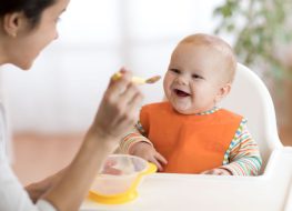 When do babies start to feed themselves with a spoon? - Mother using a baby spoon and bowl to feed her baby