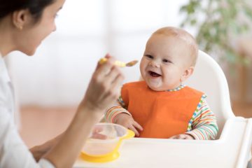When do babies start to feed themselves with a spoon? - Mother using a baby spoon and bowl to feed her baby
