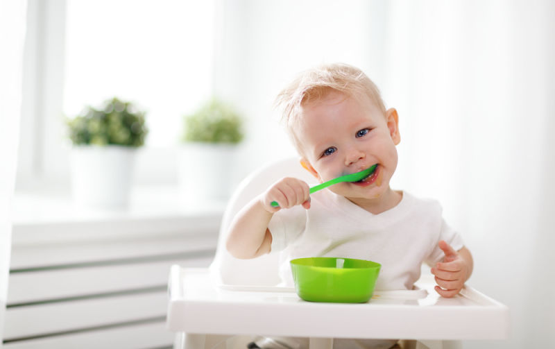 A baby feeding himself with a baby bowl and spoon.