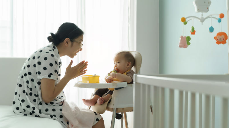 A mother feeds her baby solid foods in a high chair.