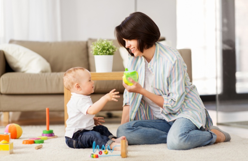 shot of a mother is showing a green and yellow sippy cup to her child while sitting in front of a sofa