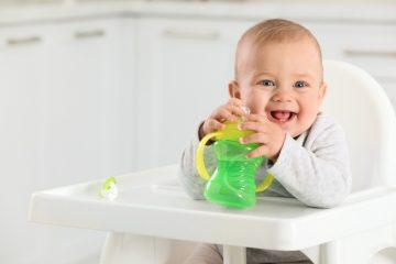 when to introduce sippy cup - shot of a baby smiling holding a green and yellow sippy cup while sitting on a white high chair