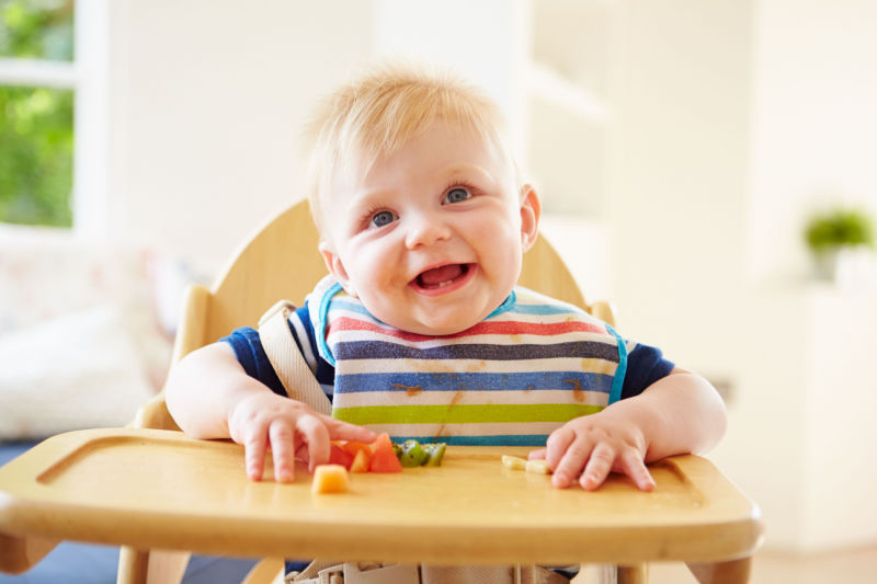 Smiling baby plays with vegetables on toddler table.