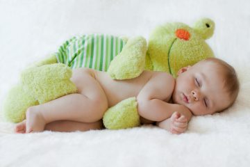a baby sleeping on his side with a stuffed frog toy cuddling him