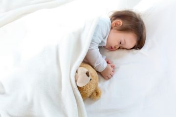 A baby sleeping on a white bed with a white blanket with a brown teddy bear beside her